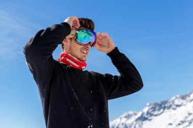 Yakışıklı bir kayakçının portresi kayak gözlüklerini ayarladı, bir kenara baktı, izole bir şekilde gökyüzünde ve tepe dağın arka planında.