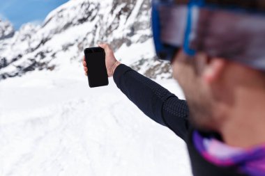 Yatay görüş. Kış sporu kıyafetleri giymiş bir snowboardcunun karlı dağlarda cep telefonuyla selfie çekerken görüntüsü. Boşluğu kopyala.