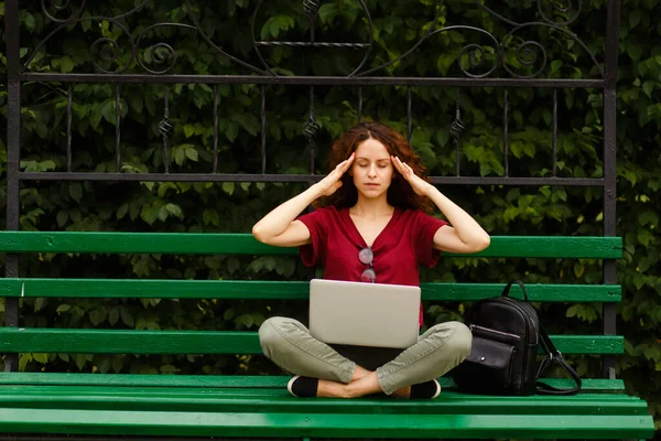 Gözleri kapalı kıvırcık bir kadın, dizüstü bilgisayarla çalışıyor, yeşil bir bankta oturuyor şakaklarına dokunuyor.. — Stok fotoğraf