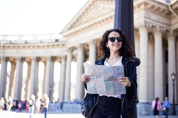 Usmívající se cestovatel žena nosí batoh a černé sluneční brýle držet mapu polohy v rukou, v blízkosti katedrály pozadí. — Stock fotografie
