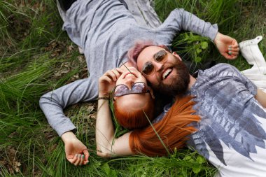 Yeşil çimenlere oturmuş kameraya bakan iki gülümseyen insanın üst görüntüsü. Yatay görünüm.