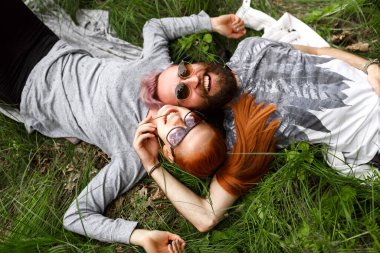 Güneş gözlüklü, yeşil çimenlere oturmuş kameraya bakan iki gülümseyen insanın en üst görüntüsü..