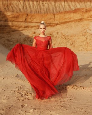 Kırmızı dalgalı elbiseli, altın rengi kumların arkasında uçan kumaşlı genç bir kadın resmi..