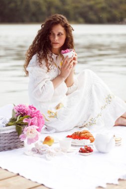 Kıvırcık saçlı bir kız, beyaz elbiseli, çay servisi yapan, piknikte kruvasan servis eden, güneşli bir zamanda, dışarıda..