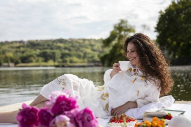 Uzun kıvırcık saçlı, beyaz elbiseli, çay servisi yapan, güneşli bir zamanda piknik yapan neşeli bir kız..