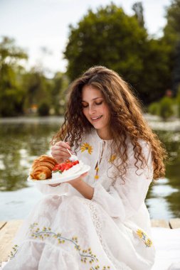 Kıvırcık saçlı mutlu kadın göl kenarında pikniğin tadını çıkar, kruvasan ye. Lezzetli ve sağlıklı bir piknik..