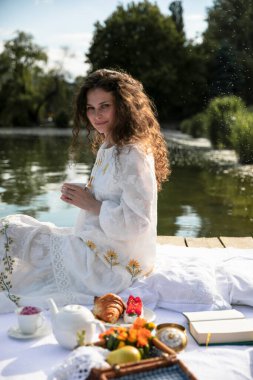 Uzun kıvırcık kızıl saçlı, beyaz elbiseli, çay servisi yapan, güneşli bir zamanda piknik yapan güzel bir kız. Dış portre.