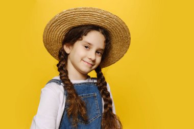 Hasır şapkalı şirin küçük kız, kameraya bakıyor, izole edilmiş sarı arka plan. Yatay görünüm.