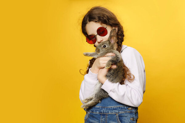 Front view of a little girl in a jeans and red sunglasses, hug her grey rabbit, isolated yellow background. Copy space.