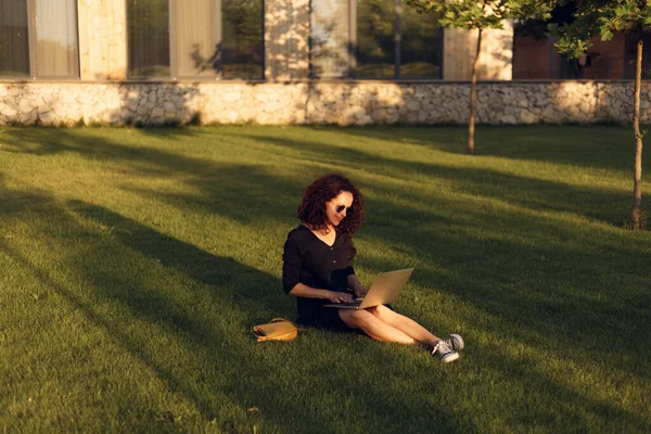 夏天的一天 戴着太阳镜的年轻漂亮的女人在草坪上坐着腿 同时还在使用笔记本电脑 在外面工作 — 图库照片
