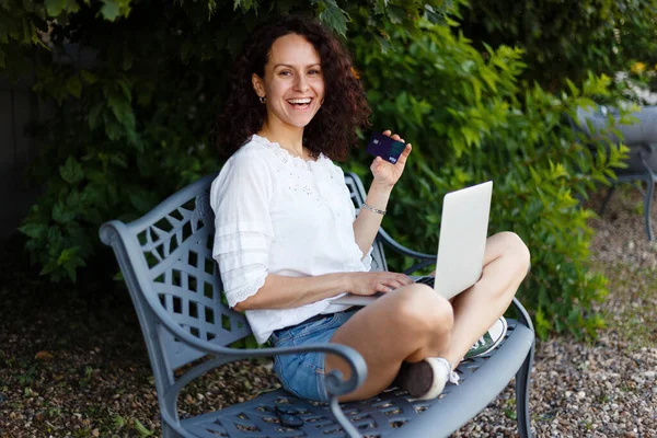 一个女孩坐在长椅上 双腿交叉在公园里 用笔记本电脑和信用卡在网上购物 — 图库照片
