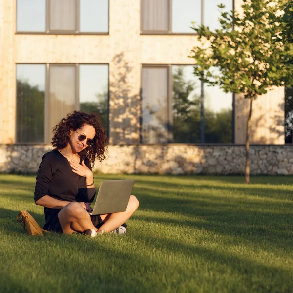 夏日里 戴着太阳镜的快乐卷曲的年轻女性坐在绿草上 双腿交叉 一边用笔记本电脑一边聊天 — 图库照片