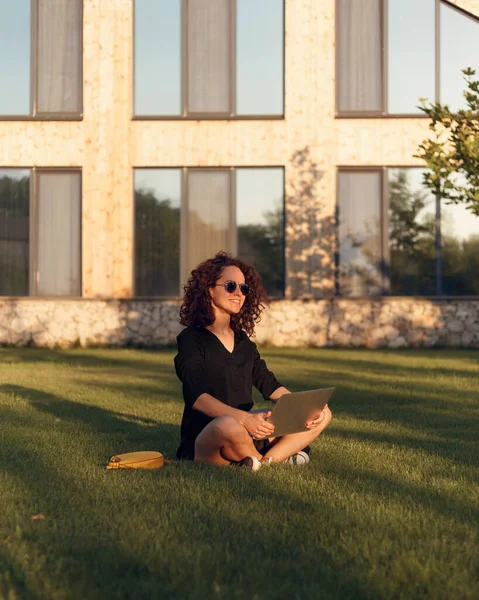 年轻美丽的女性 戴着太阳镜 坐在草地上 双腿交叉 白天使用笔记本电脑 — 图库照片