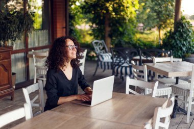 Kıvırcık saçlı, masada oturan ve terasta dizüstü bilgisayarda yazan güzel bir kadının yan görüntüsü..