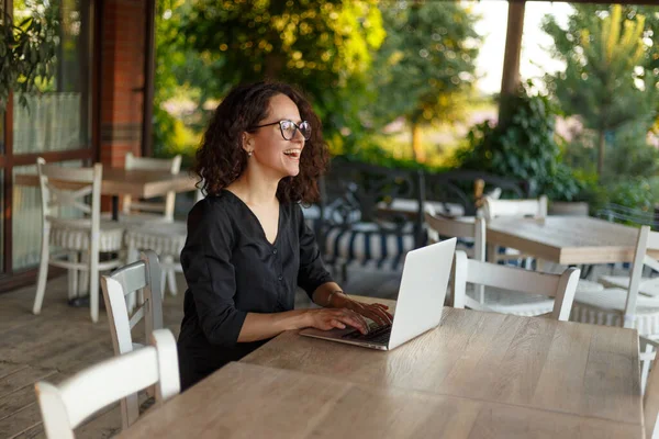 一位年轻美丽的卷发女子坐在阳台上的桌旁 在笔记本电脑上打字 — 图库照片