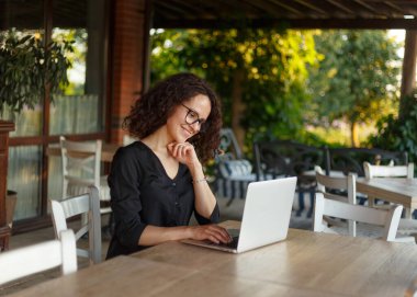 Güzel kıvırcık kadın terasta otururken dizüstü bilgisayarda çalışıyor..