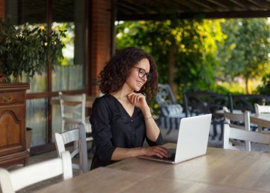 Güzel kıvırcık kadın terasta otururken dizüstü bilgisayarda çalışıyor. Yatay görünüm.