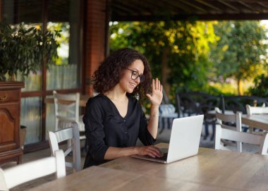 Güzel gülümseyen genç bir kadın terasta otururken dizüstü bilgisayarda çalışıyor..