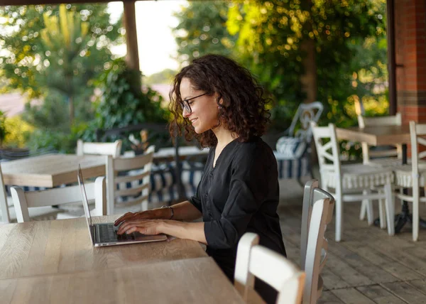 一个漂亮的年轻女子坐在桌旁 在阳台上的笔记本电脑上打字 横向观点 — 图库照片