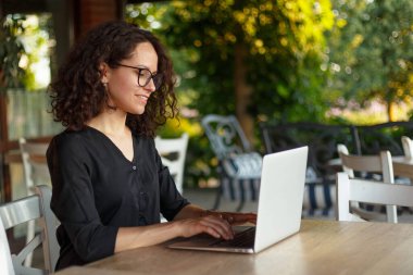 Terasta otururken dizüstü bilgisayarda çalışan genç bir kadının yan görüntüsü.
