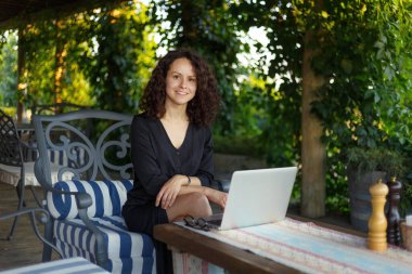 Terasta oturmuş kameraya bakarken dizüstü bilgisayarda çalışan neşeli genç bir kadın. Yatay görünüm.