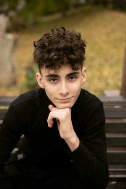 Siyah elbiseli yakışıklı bir erkek model modern saç kesimli bir bankta oturuyor ve kameraya bakıyor..