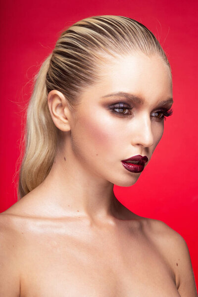 Front portrait of young female model with perfect makeup with bare shoulders, isolated on a red background.