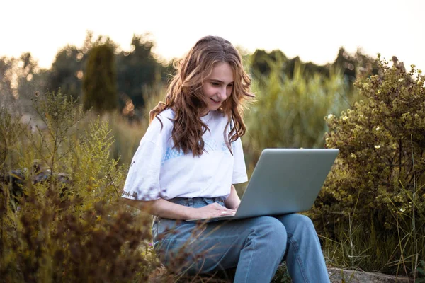 横向的观点 美丽的金发女子躺在公园的绿草上 带着笔记本电脑 面带微笑 女学生看着电脑屏幕 — 图库照片