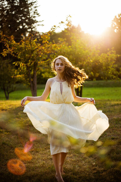 Female teens model with blonde and curly hair, turns, wear in a light dress and light in color, on the sunset background in the park