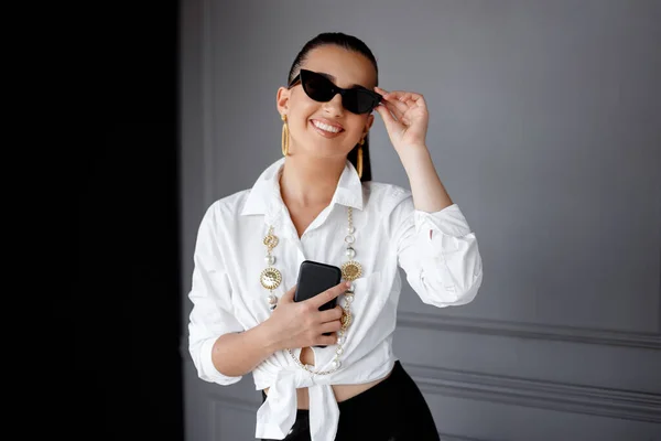 Linda jovem elegante com óculos de sol na camisa branca, segurando na mão telefone celular no fundo preto e cinza. — Fotografia de Stock
