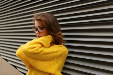 Sarı kazak giymiş, gözlük takmış, makyajlı, rüzgarda, metal duvarın yanında poz veren genç bir kadının portresi..