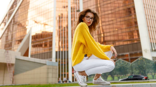 Mujer joven pensativa con el pelo ondulado, usar en suéter amarillo y jeans blancos sentados detrás de un edificio moderno. —  Fotos de Stock