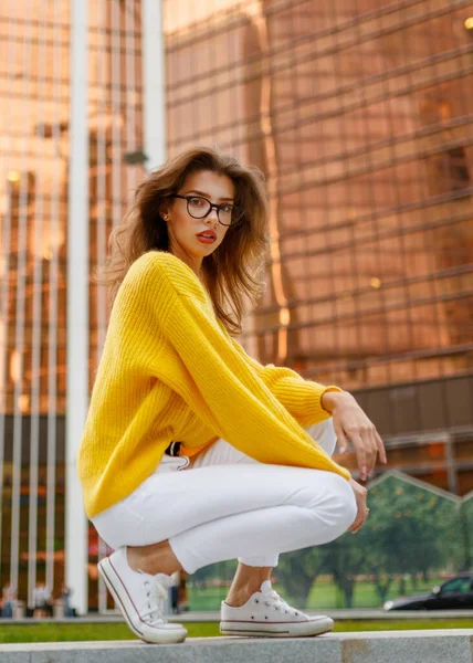 Imagen frontal de una mujer joven con el pelo ondulado, llevar en suéter amarillo y jeans blancos sentados detrás de un edificio moderno. —  Fotos de Stock