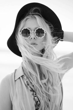 Yuvarlak gözlüklü, büyük dudaklı, dalgalı saçlı ve bordo şapkalı çekici bir sarışının ön portresi. Siyah ve beyaz.