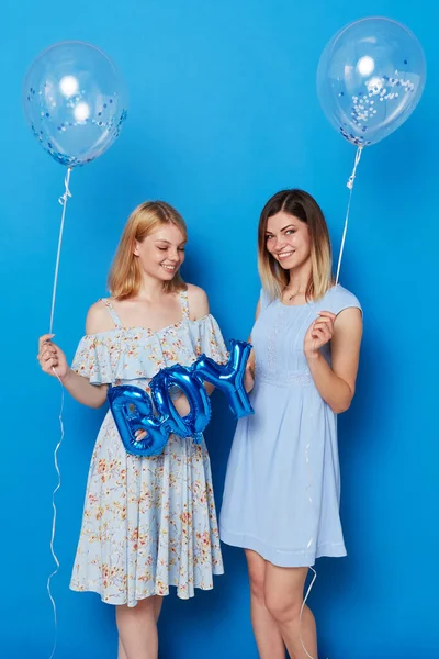 Two happy girls, young and elegant, in a studio, wearing dresses, holding blue balloons and balloon with the inscription boy, isolated blue background.