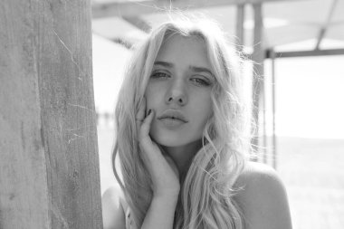 Sarışın, kıvırcık saçlı, ahşap bir terasta şehvetli poz veren ve kameraya bakan bir kadının yakın çekimi..