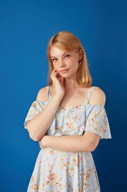 Güzel, neşeli, sarışın, sıradan giyinmiş, kameraya bakan, izole edilmiş mavi arka planı olan bir kadın..