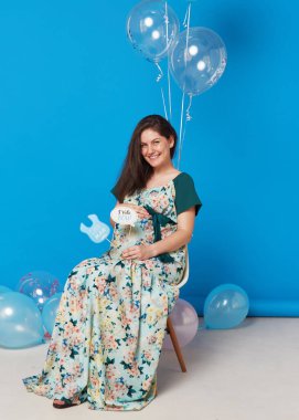 Full length image of a pregnant cheerful brunette young woman sitting on the chair with balloons, on blue background.