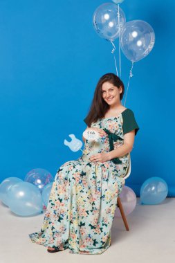 Pregnant cheerful brunette young woman sitting on the chair with balloons, on blue background.