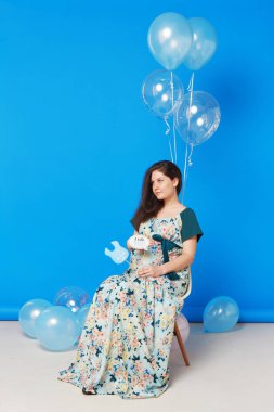 Mavi arka planda izole edilmiş balonlarla sandalyede oturan hamile esmer bir kadının tam boy görüntüsü..