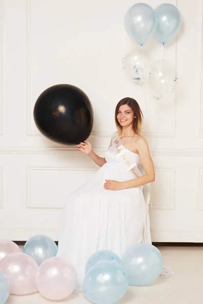 A young pregnant woman in anticipation of a baby in a white dress, hugs belly, isolated white neoclassical wall. Easy, happy maternity pregnancy, surrogacy.