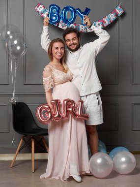 Gülen bir çiftin cinsiyet belirleme partisinde yazıtları elinde tutması, balonların üstünde parti verdiklerini gösteriyor..