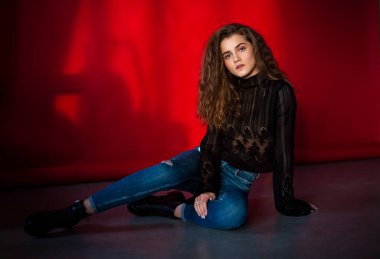 Makyajlı, kıvırcık saçlı, siyah bluz ve kot pantolonlu, izole kırmızı arka planı olan çekici genç bir kadın..