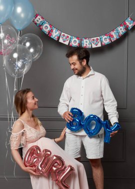 Cinsiyet belirleme partisinde balon tutan heyecanlı genç bir kadın ve erkek resmi. Stüdyoda, gri duvarın yanında poz veriyor..