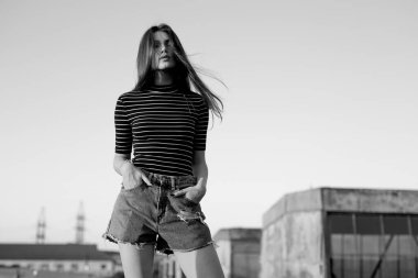 Black and white picture. Young hipster woman posing sensually on the roof, her hair is in wind at sunset.