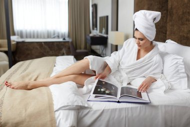 Cheerful young woman in white bathrobes with towel on hier head, browse a magazines, lying down on bed.