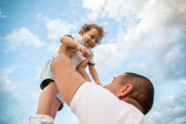 Baba, çocuğu mavi gökyüzüne doğru fırlatıyor. Mutlu çocuk babasıyla yeşil bir alanda ve mavi gökyüzünde oynuyor.