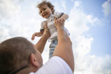 Baba, çocuğu mavi gökyüzüne doğru fırlatıyor. Mutlu çocuk mavi gökyüzünün arka planında babasıyla oynuyor.