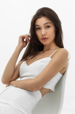 Makyajlı ve saç stili beyaz giyinmiş esmer kadın koltuğa rahatça oturur.