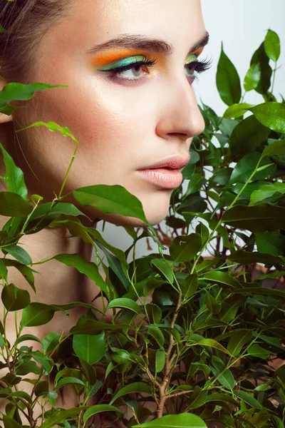 Young beautiful woman with healthy skin of face and palm leaves. Closeup fresh face of an attractive caucasian girl with green plants. Model with bright brown eye makeup.
